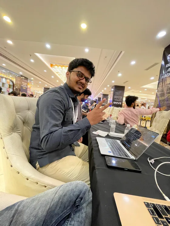Mirza in a tufted white chair at his laptop, making a peace sign