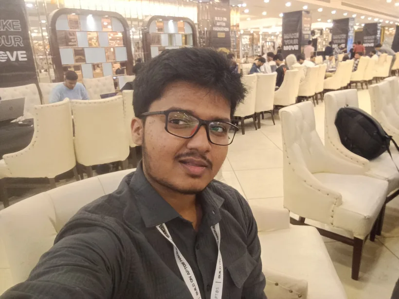 A selfie of Mirza in the hall, with rows of white chairs and hackers behind him
