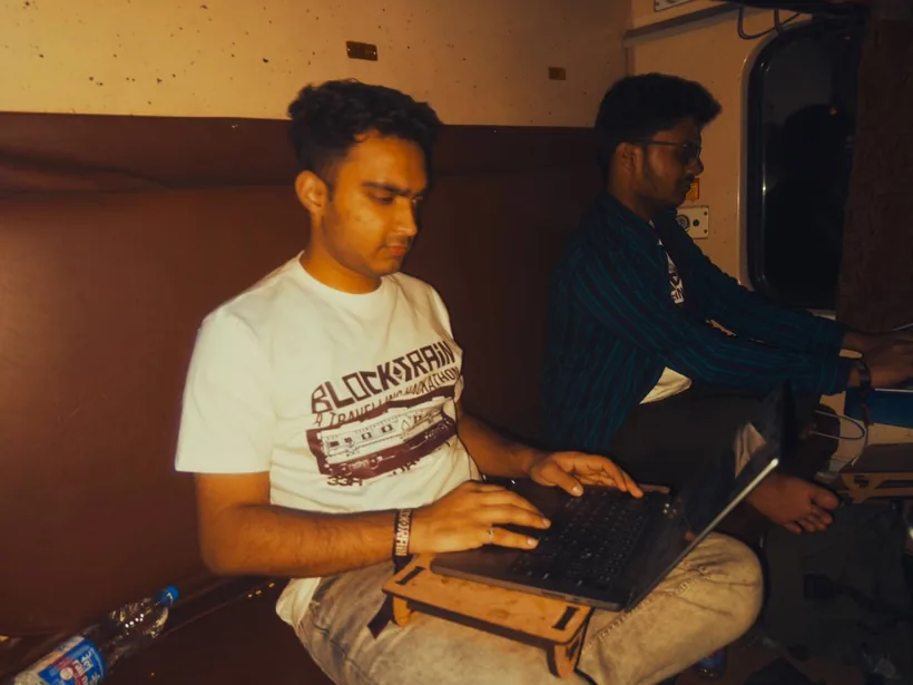 A hacker typing on a laptop in a dim train compartment while another works behind him