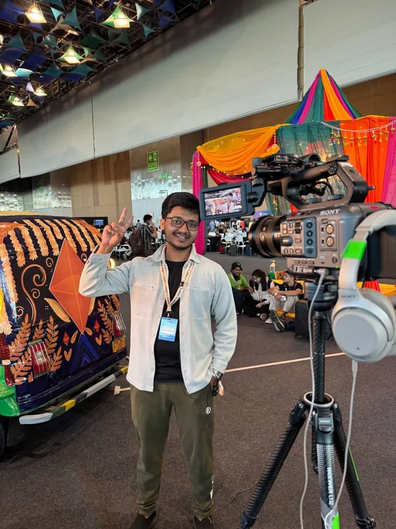 Mirza flashing a peace sign next to a painted auto rickshaw, with a video camera in the foreground