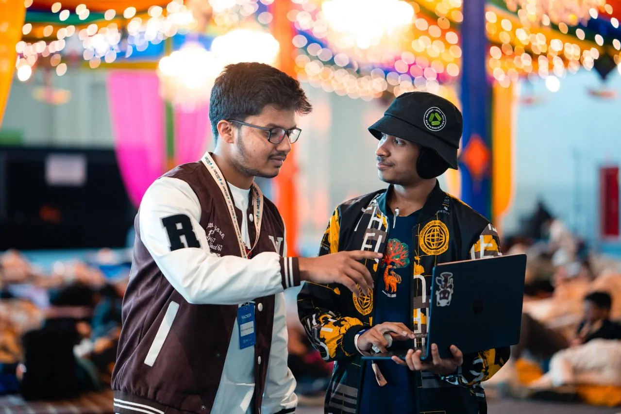 Mirza pointing at a laptop screen while a teammate in a bucket hat holds it, under strings of lights at the venue
