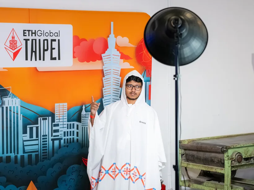 Mirza in a white rain poncho, pointing up, in front of the orange ETHGlobal Taipei backdrop with Taipei 101